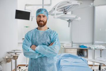 Smiling male surgeon standing in operating room with crossing hands, ready to work on patient