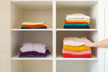 Young adult woman hand sorting folded colorful different shirt, jeans, sweaters and sport hoodie jackets on shelves in white opened wardrobe at room. Front view. Closeup. Female casual clothes.
