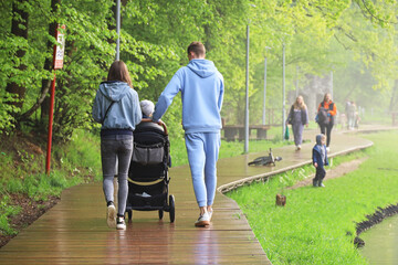 Young couple with baby pram walking in a park after rain. Family leisure on lake coast in summer