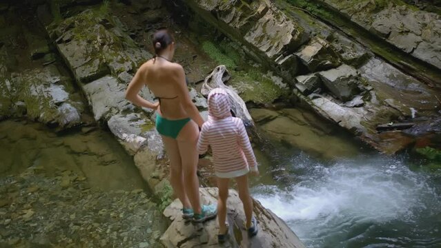 Woman with child at waterfall in forest. Creative. Woman in swimsuit and boy are standing at waterfall in forest. Family hike with swimming in waterfall in wild forest
