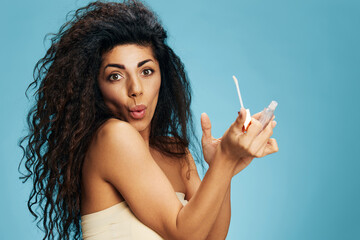 Obraz premium Shocked excited attractive curly Latin lady hold lip gloss posing isolated over pastel blue background. Cosmetic lips product ad Natural beauty Makeup sensation concept Studio portrait Copy space