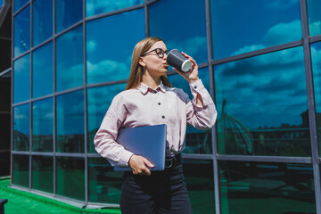 A woman in glasses with coffee in her hands, against a skyscraper. Modern building, financial center, cityscape. Successful female entrepreneur with glasses