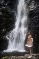 Attractive girl in a beige dress is posing on the background of the waterfall.