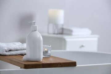 Fototapeta na wymiar Bottle of bubble bath with foam, towel and candle on tub in bathroom, space for text