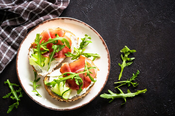 Open sandwich with cream cheese and prosciutto or jamon with fresh greens in white plate. Top view at dark table.