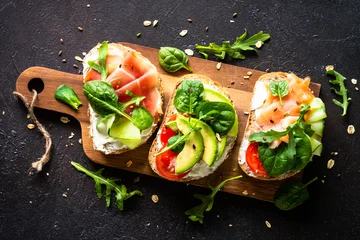 Fototapeten Open sandwich set with cream cheese, prosciutto, salmon, avocado and fresh greens. Top view at dark. © nadianb