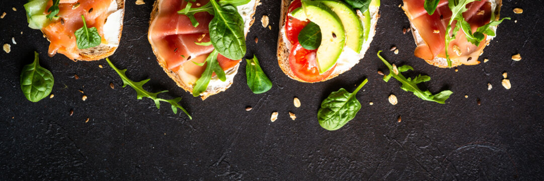 Open sandwiches set with cream cheese, prosciutto, salmon, avocado and fresh greens and vegetables. Long banner format.