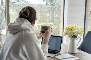 Back view of middle-aged woman with coffee mug working from home office. Freelancer taking a coffee break.