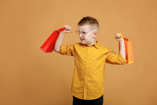 Image of smiling boy holding bags with presents or shoppings on yellow background. child in glasses lifts packages like weights