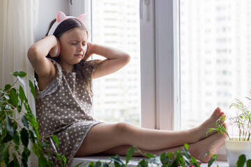 Fototapeta na wymiar Cute little girl with wireless headphones sitting near window at home.