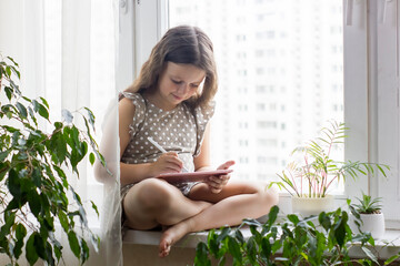 Cute little girl sitting near window at home and drawing with a digital tablet and a pen stylus.