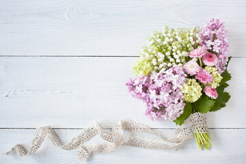 Spring bouquet of lilies of the valley, pink lilacs, daisies, viburnum flowers with lace ribbon on a white wooden background, space for text.