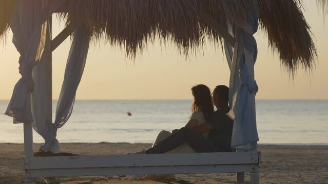 Couple of lovers sit on canopy, man and woman embraces admire serene sea