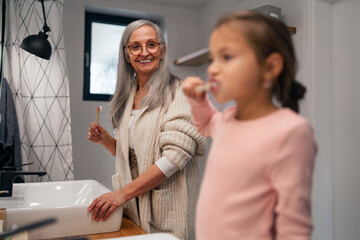 Senior grandmother making a ponytail to her granddaughter indoors in bathroom in the morning.