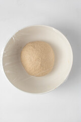 Tightly rolled ball of dough in a white bowl covered with clingfilm, ball of pastry dough left to rise