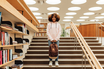 Schoolgirl with backpack standing on stairs in library. Smiling student in casuals looking at...