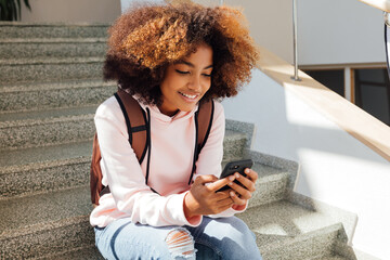 Portrait of a smiling girl in casuals sitting on stairs in school holding smartphone