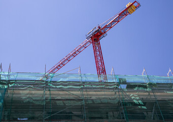 Fototapeta na wymiar Red construction crane against blue sky at construction site. A facade of an old building closed for renovation, repair with green protective construction mesh. Engineering architectural business.