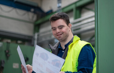 Young man with Down syndrome looking at blueprints when working in industrial factory, social...