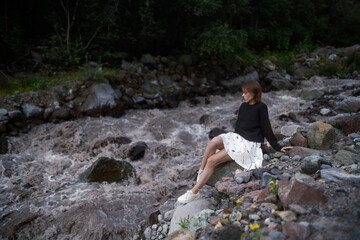 Portrait of an elegant woman in black and white clothes in nature as advertising cover or promotion design. Cute brunette woman sits on the stones next to the river. in nature. 