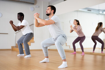 Young bearded man doing squats during warm up before group yoga class in fitness room
