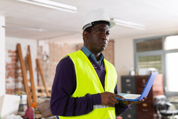 Portrait of african-american man builder in vest standing inside apartment during repair works with laptop.