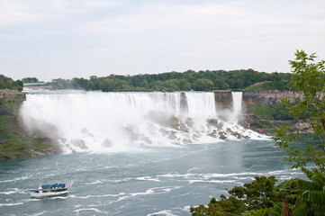 The small Maid of the Mist below the iconic tourist attraction Niagara Falls with its dramatic drop.