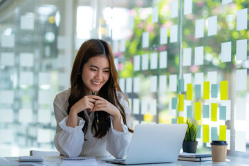 Happy Asian businesswoman laughing sitting at the work desk with laptop, a cheerful smiling female employee having fun feeling joy and positive emotion express sincere laughter at office workplace