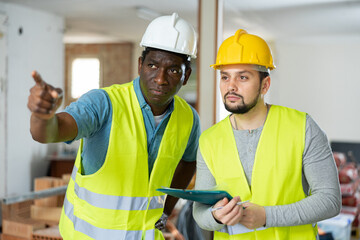 Two builders working on a construction site indoors are taking important notes on a tablet,...
