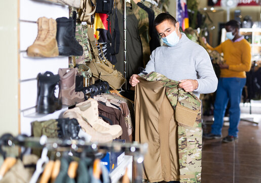 European man in face mask choosing camouflage clothes in military store. African-american man shopping in background.