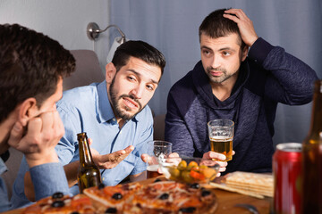 Troubled thoughtful male friends talking, drinking beer with pizza