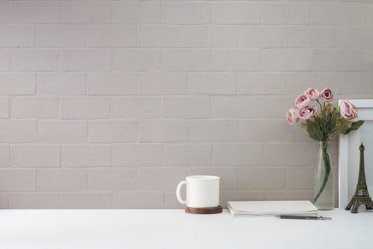 Feminine workplace with picture frame, coffee cup, book and flower pot on white table against brick wall. Copy space for your text.