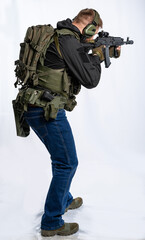Fototapeta premium an airsoft soldier in full gear with his back to us is aiming at an optical sight. a man in an outfit, in headphones, a bulletproof vest, with a backpack and a belt. White background.