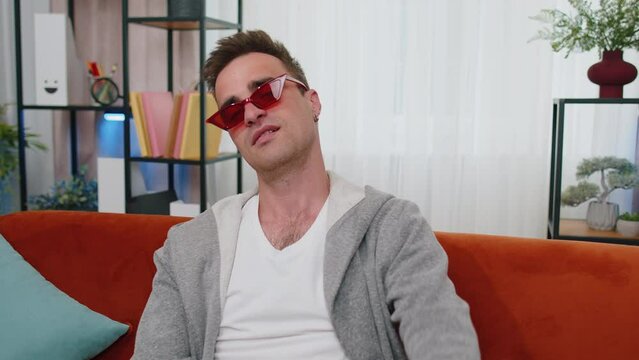 Portrait of seductive cheerful stylish successful man in t-shirt wearing red sunglasses, charming smile, blinking, winking eye, flirting. Young lovely adult guy sitting at home in living room on couch