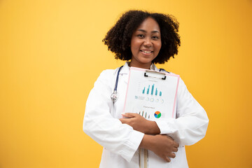 Africa American beautiful charming doctor wearing white gown uniform with stethoscope, holding clip board of health graph and finance report, happily smiling, standing on isolated yellow background