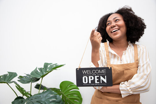 Portrait of a happy waitress standing in white restaurant with monstera. Happy woman owner showing open sign in her small business shop. Positive thinking and charming