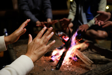 Fototapeta na wymiar Close-up on hand of woman warming on campfire. Group of asian friends sitting around campfire in the night. Friends camping near bonfire relaxing on winter vacation at the night time.