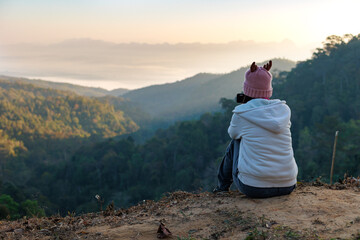 Naklejka premium Rear view of asian woman sitting on the ground looking on the view of beautiful nature on the top of mountain. People enjoying with panoramic view in the morning time.