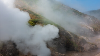 Clouds of steam from an erupting geyser envelop the hillsides. Poor visibility due to haze. Blue sky, green slope. Kamchatka. Valley of Geysers