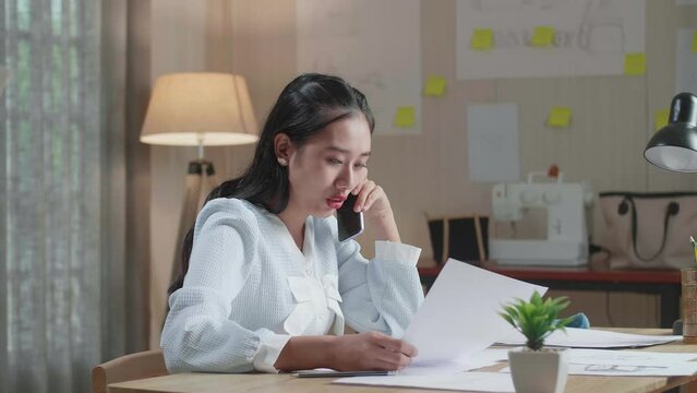 Asian Woman Designer Talking On Smartphone While Looking At The Layout Bond In Hand At The Office
