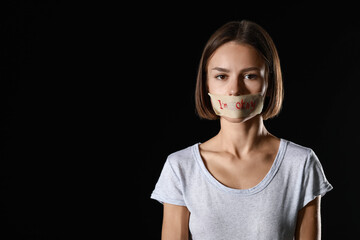 Scared young woman with taped mouth on black background. Violence concept