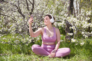 Naklejka na ściany i meble Spring awakening. Slow life. Enjoying the little things. Dreaming of spring. Middle aged woman in sportswear and headphones uses smartphone in a spring flowering garden. social networks