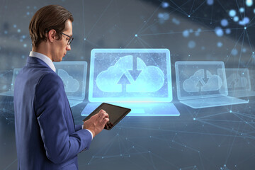 Attractive young caucasian businessman with tablet and abstract polygonal digital laptop with cloud computing hologram on blurry interior background. Database and information concept. Double exposure.