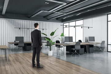 Fototapeta premium Attractive thoughtful young european man standing in wooden and concrete office interior with furnituire and window with city view. CEO, executive and consulting concept.
