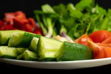 Close-up of chopped fresh herbs and vegetables on a white plate on a black background. Cucumbers, tomatoes, bell peppers, green onions, cockerel. Useful vitamins from vegetables