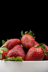 Close up ripe red strawberries with green tails in a white plate on a black background. Sweet dessert of fresh berries on the table. Vertical photo