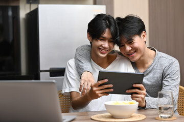 Young gay couple using digital tablet on bed at home, having fun checking social media applications together