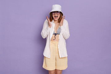 Naklejka premium Portrait of delighted young Caucasian woman wearing swimsuit, shirt, shorts and panama posing isolated over purple background, being surprised, raised arms, screaming happily.