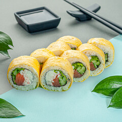 Sushi roll with salmon wrapped in egg omelet. With cucumber crab and cream cheese. Decorated with greenery. In the background is a gravy boat and chopsticks. Close-up.