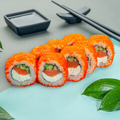 Sushi roll with salmon, butter cream and cucumber. Sprinkled with masago caviar. Decorated with greenery. In the background is a gravy boat and chopsticks. Close-up.